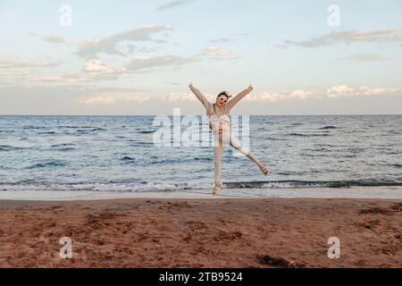 Junge glückliche Frau in elfenbeinfarbenem Hoodie und Leggings und weißer Sonnenbrille, die am Strand spreizt. Wunderschönes Naturkonzept von Wasser und Sand Stockfoto