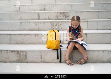 Schulmädchen, Rucksack in der Nähe, konzentriert sich auf ihre Hausaufgaben, schreibt Notizen auf dem Schulhof. Stockfoto