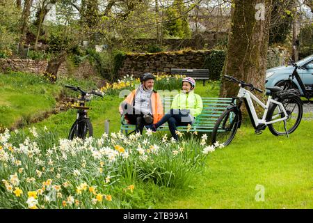 Mann und Frau Radfahrer mit Elektro-E-Bikes sitzen im Frühling auf einer Bank in einem hübschen Dorf, Hartley. Kirkby Stephen, North Pennines, Cumbria Stockfoto