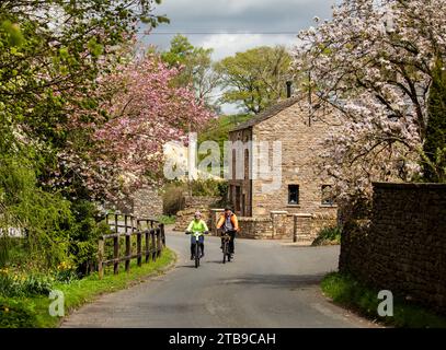 Mann und Frau Radfahrer fahren E-Bikes in einem hübschen Dorf im Frühling, Hartley. Kirkby Stephen, North Pennines, Cumbria Stockfoto