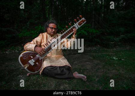 Der Hindu-Mann sitzt am Rande eines Waldes und spielt die Sitar in einem Sivananda Yoga Asram in Quebec, Kanada Stockfoto