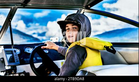 Porträt eines Jungen mit Schwimmweste und Kapuze, der am Lenkrad eines Motorbootes auf dem Shuswap Lake sitzt; British Columbia, Kanada Stockfoto