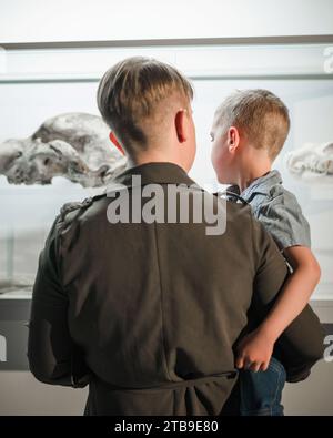 Neugieriger kleiner Junge in den Armen seiner Mutter, der ein Tierkelett in einer Knochenvitrine in einer Naturkundeausstellung ansieht. Stockfoto