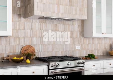 Ein Küchenofen und eine Kapuze mit braunen rechteckigen Fliesen, Edelstahlofen, weißen Schränken und gemütlichem Dekor auf Arbeitsflächen aus grauem Stein. Stockfoto