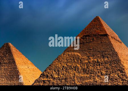 Zwei der wichtigsten Pyramiden von Gizeh, sonnenbeleuchtet mit einem goldenen Farbton, die große Pyramide (Khufu) unter einem dunklen, bewölkten Himmel; Gizeh, Ägypten Stockfoto