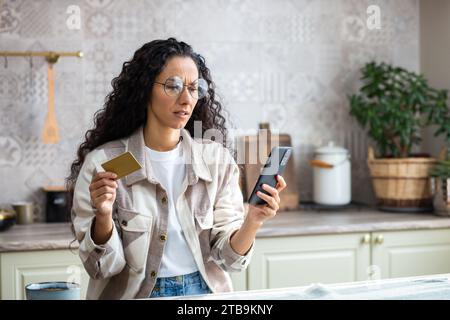 Frustrierte betrogene Frau zu Hause in der Küche abgelehnt, falscher Geldtransfer, Internetbetrug, lateinamerikanische Frau traurig, die Bank Kreditkarte und Telefon hält, die Online-Shopping-App benutzt. Stockfoto