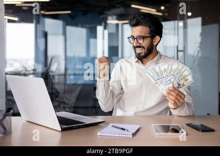 Junger indischer Mann, der im Büro am Laptop arbeitet, Geld in der Hand hält, glücklich mit Erfolg, Siegesgesgestik mit der Hand zeigt. Stockfoto