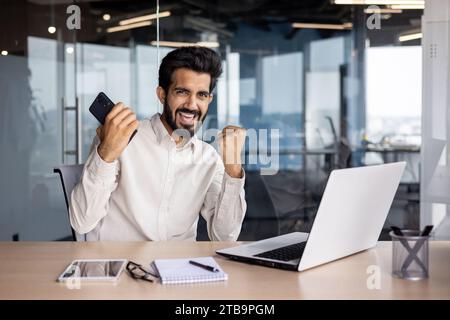 Porträt eines jungen indischen Geschäftsmannes, freiberuflich im Büro am Tisch sitzt, das Telefon in den Händen hält und in die Kamera blickt, mit den Händen eine Siegesgesgestik zeigt. Stockfoto