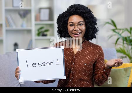 Porträt einer lächelnden afroamerikanischen Lehrerin, die zu Hause auf dem Sofa sitzt und ferngesteuert unterrichtet. Sie hält eine Tafel mit einer Lektion in der Hand. Nahaufnahme des Fotos. Stockfoto