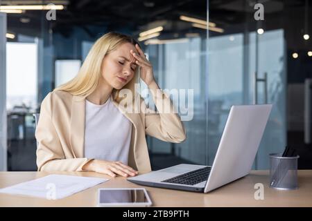 Müde und überarbeitete junge ukrainische Frau arbeitet im Büro am Laptop, hält den Kopf, fühlt starke Schmerzen und Druck. Stockfoto
