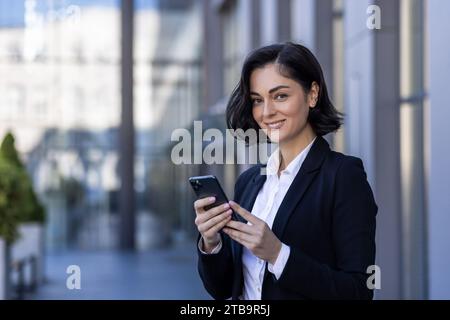 Nahaufnahme eines Porträts einer jungen Geschäftsfrau, die in der Nähe eines Bürogebäudes steht, ein Telefon hält und in die Kamera lächelt. Stockfoto