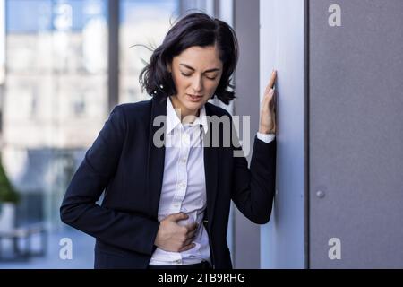 Eine junge Geschäftsfrau mit Bauchschmerzen, die neben dem Bürogebäude steht, sich an die Wand lehnt und ihren Körper hält. Stockfoto