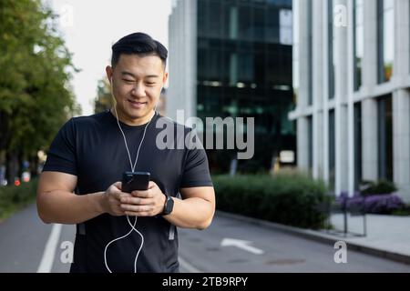 Ein lächelnder junger asiatischer Mann, der morgens mit Telefon und Kopfhörern joggt. Schaltet Musik und Podcast ein. Stockfoto