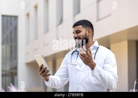 Ein junger muslimischer männlicher Arzt steht in einem weißen Mantel neben dem Krankenhaus auf der Straße und spricht auf dem Anruf mit Kollegen, ein Patient mit einer Tablette, berät, mit den Händen Gesten. Stockfoto