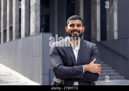 Porträt eines lächelnden indischen Geschäftsmannes, der in einem Anzug auf der Straße in der Nähe des Büros steht, seine Arme kreuzt und die Kamera anlächelt. Nahaufnahme. Stockfoto