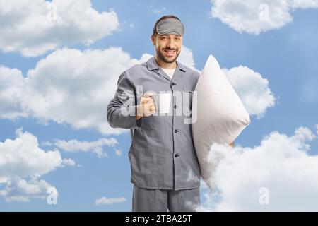 Ein Mann im Pyjama und einer Schlafmaske, der eine Tasse Kaffee vor Wolken auf einem blauen Himmel hält Stockfoto