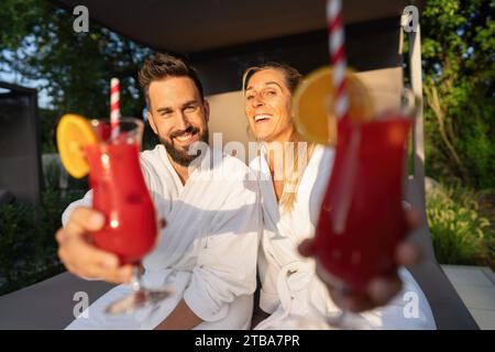 Fröhliches Paar in weißen Spa-Bademänteln, das rote Cocktails in einem Wellness-Resort genießt Stockfoto