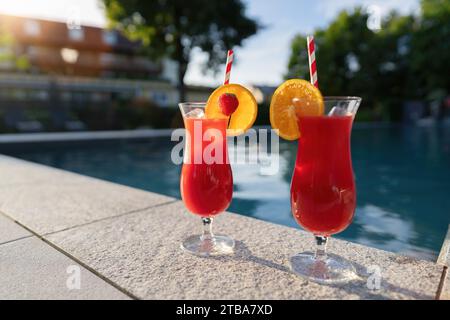 Zwei rote Cocktails mit Obstgarnituren am Pool, die das warme Licht des Sonnenuntergangs reflektieren Stockfoto