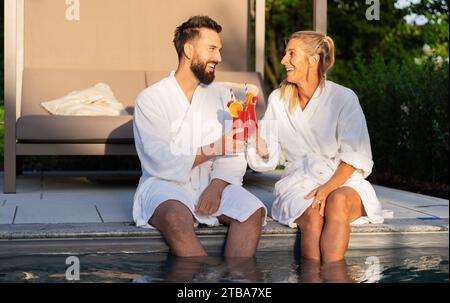 Zwei Personen in weißen Bademänteln lachen und toasten mit roten Getränken am Pool bei Sonnenuntergang in einem Wellnesshotel Stockfoto