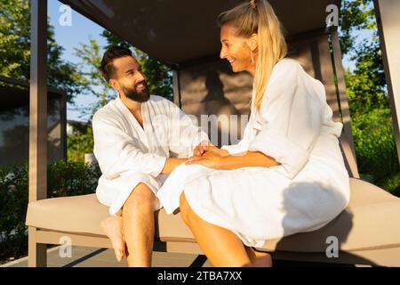 Ein lächelndes Paar verliebt in weiße Bademäntel, das im Sonnenlicht auf einer Sitzbank im Freien im Spa Wellness Hotel sitzt Stockfoto