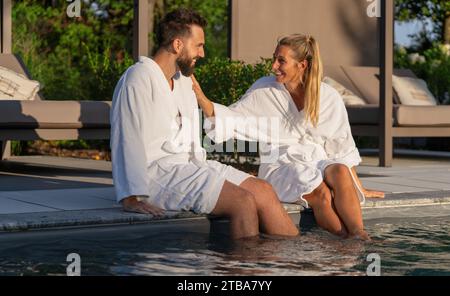 Paare in weißen Bademänteln sitzen mit Füßen im Pool und genießen einen sonnigen Tag im Spa Wellness Hotel Stockfoto