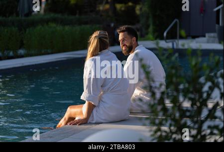 Ein Paar in weißen Bademänteln, das sich hinten an der Kante eines Pools in einem Wellnesshotel befindet Stockfoto