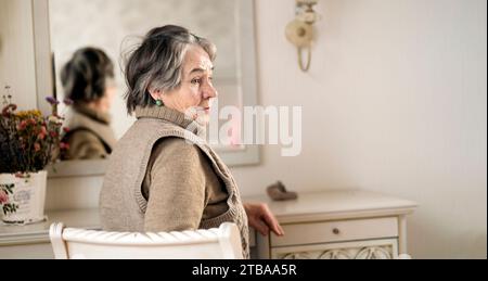 Schöne alte Frau sitzt neben dem Spiegel in ihrem gemütlichen hellen Raum. Stockfoto