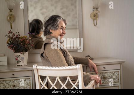 Schöne alte Frau sitzt neben dem Spiegel in ihrem gemütlichen hellen Raum. Stockfoto