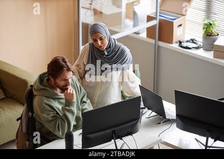 Hochwinkelporträt einer muslimischen jungen Frau, die mit einem männlichen Kollegen im Amt auf den Computerbildschirm blickt Stockfoto
