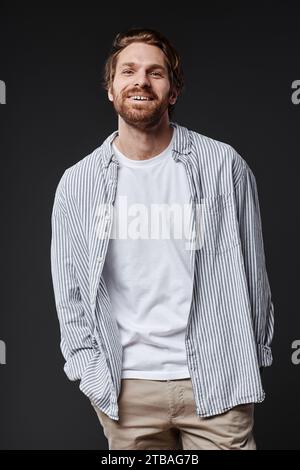 Vertikales Porträt eines lächelnden bärtigen Mannes, der im Atelier mit sorglosem Gesichtsausdruck in die Kamera blickt Stockfoto