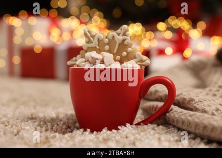 Köstlicher Kakao mit Marshmallows und Lebkuchenkeks in der Tasse auf weichem Teppich vor verschwommenem Weihnachtslicht, Nahaufnahme Stockfoto