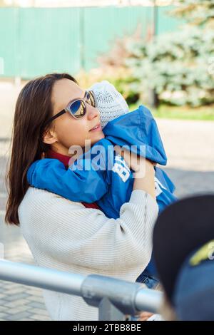 Die fürsorgliche Mutter tröstet ein kleines Kind, das auf der Straße weint. Die Frau trägt ein verärgertes Baby, das auf dem Stadtplatz läuft. Lady beruhigt das Kind Stockfoto