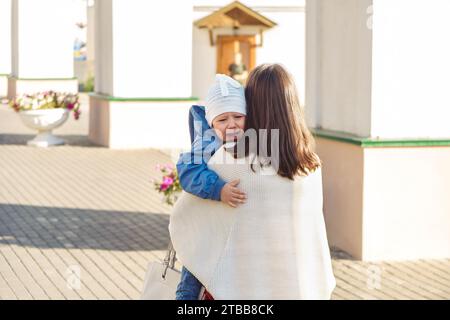 Die fürsorgliche Mutter tröstet ein kleines Kind, das auf der Straße weint. Die Frau trägt ein verärgertes Baby, das auf dem Stadtplatz läuft. Lady beruhigt das Kind Stockfoto