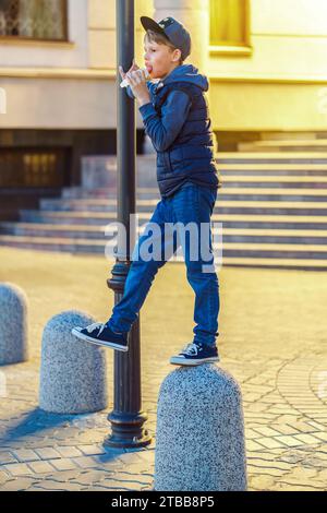 Ein verspielter Junge isst Eis auf Steinsäule in der Abendstadt. Das Kind leckt Eisbecher im Kegel und hat Spaß auf der Straße. Urbanes Lifestyle und Street Food Stockfoto