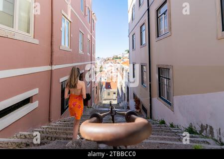 Hochwinkelansicht einer jungen Frau in einem orangefarbenen Kleid, die steile Steintreppen in Lissabon, Portugal hinabsteigt, horizontal von hinten und oben aufgenommen, Stockfoto