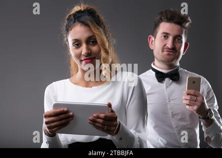 Eine junge schwarze Frau hält ein Tablet, ein weißer Mann mit Telefon im Hintergrund Stockfoto