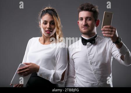 Elegant gekleidete Frau konzentriert sich auf ein Tablet, Mann überprüft sein Handy Stockfoto