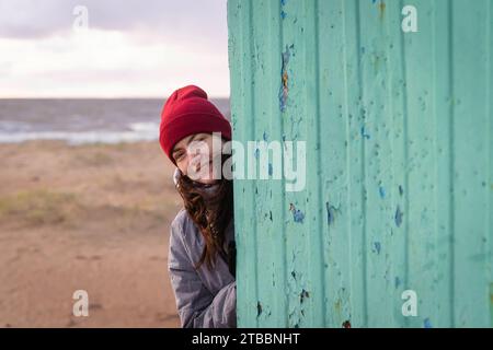 Ein friedlicher Herbsttag am Strand, während eine Frau in einem stilvollen roten Hut anmutig vor einem alten Umkleidestand posiert und eine ruhige und gemütliche Atmosphäre schafft Stockfoto