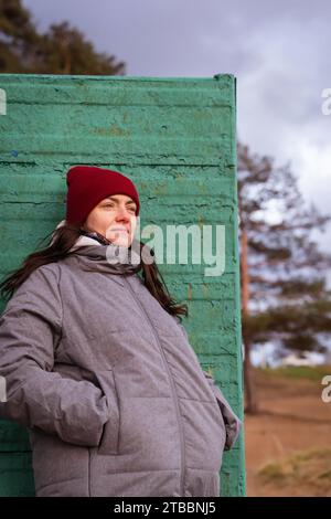 Eine modische junge Frau mit einem knallroten Hut schlendert an einem malerischen Herbststrand entlang und posiert neben einem charmanten alten Umkleidestand Stockfoto