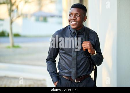 Junger schwarzer Geschäftsmann, der vor dem Gebäude mit Tasche steht und lächelt. Stockfoto