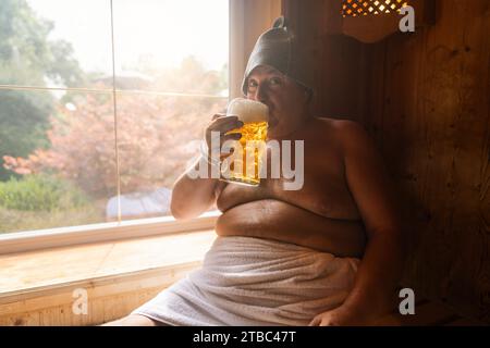 Mann in einer finnischen Sauna mit Filzmütze, schlürft an einem deutschen Bierbecher, mit einem hellen Fenster im Hintergrund. Oktoberfest-Konzeptbild Stockfoto