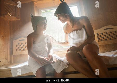 Mutter und Kind lächeln einander in einer finnischen Sauna an, beide tragen Filzhüte Stockfoto