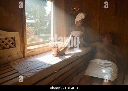Paare, die sich in der Sauna entspannen, Frau mit einem Handtuch auf dem Kopf, beide sitzen und lächeln im Wellnesshotel Stockfoto