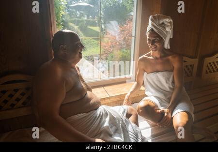 Mann und Frau in der Sauna, Frau mit einem Handtuch auf dem Kopf, beide lächeln und sitzen im Wellnesshotel Stockfoto