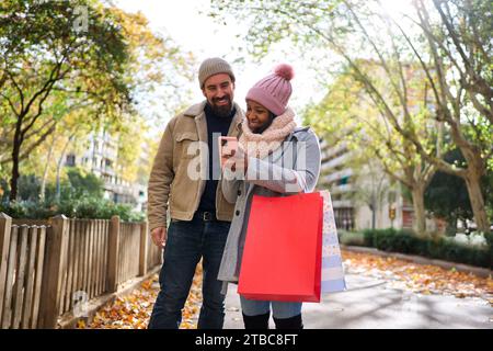 Junge interrassische Paare, die das Telefon benutzen, während sie auf der Straße spazieren gehen und gemeinsam weihnachtseinkäufe machen. Stockfoto