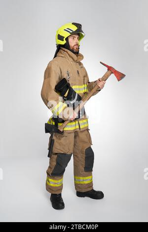 Ernsthafter bärtiger Feuerwehrmann in voller Ausrüstung mit roter Axt für die Brandbekämpfung. Seitenansicht eines fokussierten männlichen Retters mit Beil-Feuerwerkzeug, auf grauem Atelierhintergrund. Konzept der Arbeit, Brandausrüstung, Sicherheit. Stockfoto