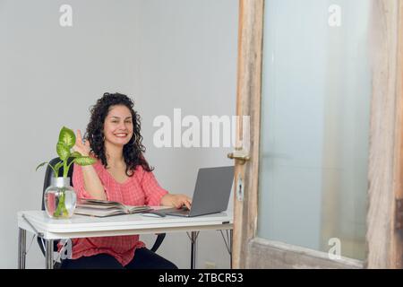 Junge glückliche Lateinfrau mit Locken, sitzt zu Hause in ihrem neuen Büro, lächelt und blickt in die Kamera, sehr glücklich bereit, an ihrem Abenteuer zu arbeiten. Stockfoto