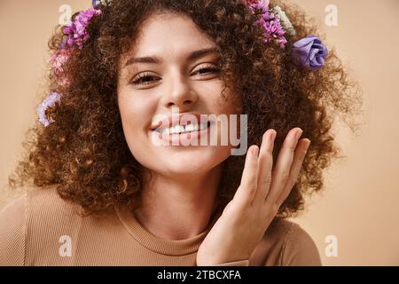 Porträt einer glücklichen Frau mit bunten Blumen im Haar lächelnd in die Kamera auf beige, natürliche Schönheit Stockfoto