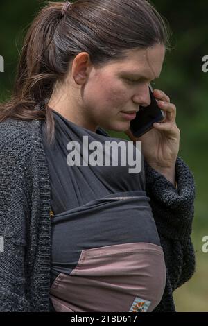 Junge Mutter am Telefon mit ihrem Smartphone, ihr Baby in eine Babytragetasche gewickelt, Mecklenburg-Vorpommern, Deutschland Stockfoto