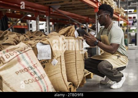 Seitenansicht Porträt des jungen Schwarzen Mannes, der ein Tablet im Lager der Kaffeerösterei benutzt und Bestandskontrolle, Kopierraum macht Stockfoto
